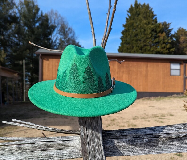 May include: A green felt fedora hat with a brown leather band and a forest scene design. The hat is sitting on a wooden fence.