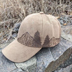 May include: A brown suede baseball cap with a brown embroidered design of mountains and trees. The cap has a curved brim and a structured crown.