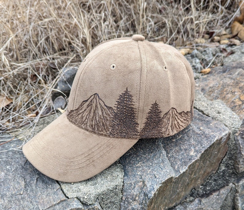 May include: A brown suede baseball cap with a brown embroidered design of mountains and trees. The cap has a curved brim and a structured crown.
