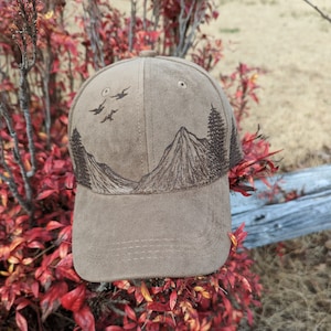 May include: A brown suede baseball cap with a mountain and birds design embroidered on the front. The cap has a mesh back and a brown adjustable strap.