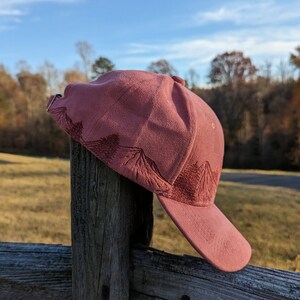May include: A pink baseball cap with a mountain design embroidered on the front. The cap is hanging on a wooden fence post.