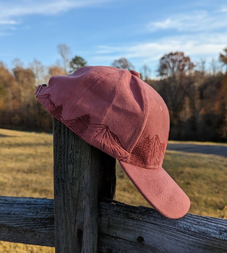 May include: A pink baseball cap with a mountain design embroidered on the front. The cap is hanging on a wooden fence post.