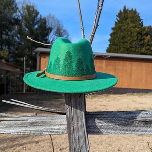 May include: A green felt fedora hat with a brown leather band and a forest print. The hat is sitting on a wooden fence.