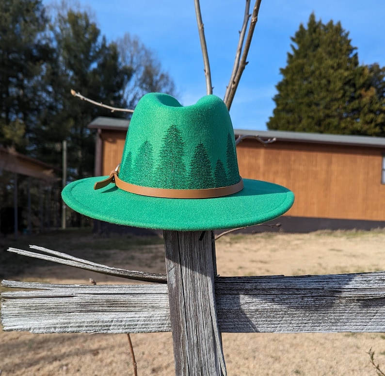May include: A green felt fedora hat with a brown leather band and a forest print. The hat is sitting on a wooden fence.