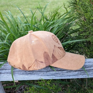 K&ouml;nnte beinhalten: Eine braune Baseballkappe mit einem braunen gestickten Design von Bergen und einem Hei&szlig;luftballon. Die Kappe hat einen gebogenen Schirm.