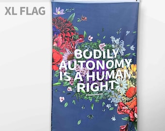 May include: A blue flag with a floral design and the text "BODILY AUTONOMY IS A HUMAN RIGHT" in white. The text is surrounded by colorful flowers and leaves. The flag is hanging from a white pole.