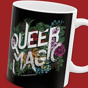 Puede incluir: Taza de cerámica blanca con fondo negro que presenta el texto "QUEER MAGIC" en letras blancas rodeado de ilustraciones florales de colores. El texto "@TRANSPAINTER" está en la parte inferior de la taza.