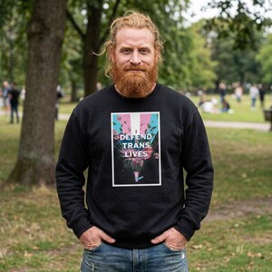 May include: Black sweatshirt featuring the text "DEFEND TRANS LIVES" in white, pink, and blue, with floral accents. The image is set in a park with trees and greenery.