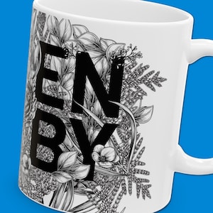 Könnte beinhalten: Weiße Keramiktasse mit schwarzem Blumendesign und dem Wort "ENBY" in schwarzen Buchstaben.