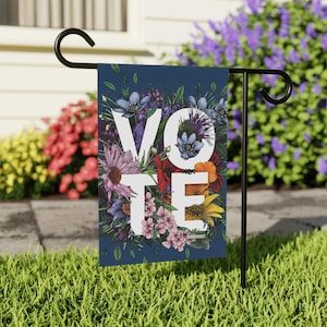 Peut inclure: Un drapeau de jardin bleu marine avec le mot "VOTE" en grandes lettres blanches. Les lettres sont entourées d'un arrangement floral coloré de fleurs violettes, roses, rouges et jaunes. Le drapeau est sur un support en métal noir.