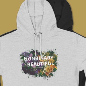 May include: A light gray hoodie with a floral design and the text "NONBINARY IS BEAUTIFUL". The design features a variety of flowers in shades of purple, yellow, and green. The hoodie has a drawstring hood.