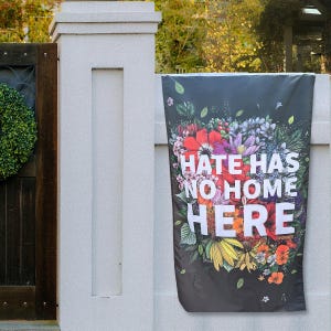 May include: A black garden flag with the text "HATE HAS NO HOME HERE" in white, surrounded by a colorful floral arrangement. The flag is hanging on a white wall next to a wooden gate.