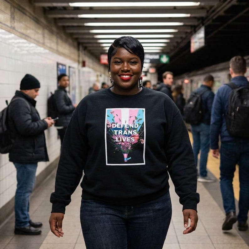 May include: A black sweatshirt with the text "DEFEND TRANS LIVES" in a white rectangle with a floral design. The person is wearing blue jeans and is in a subway station.