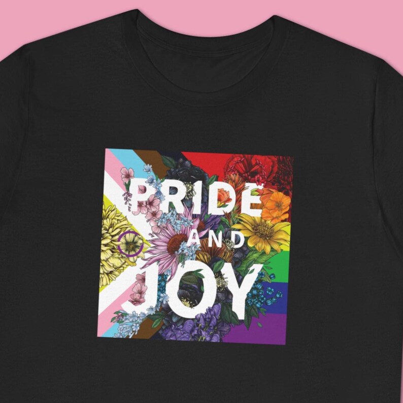 May include: Black t-shirt featuring a floral design with the words "PRIDE AND JOY" in white. The floral arrangement includes a variety of colorful flowers and the Pride flag colors, celebrating LGBTQ+ pride.