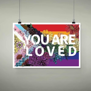 May include: A poster with the words "YOU ARE LOVED" in white, set against a rainbow background. The poster is adorned with a vibrant floral arrangement, including pink, purple, yellow, and red flowers, and the Pride flag.