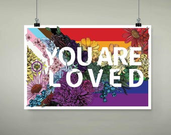 May include: A poster with the words "YOU ARE LOVED" in white, set against a rainbow background. The poster is adorned with a vibrant floral arrangement, including pink, purple, yellow, and red flowers, and the Pride flag.