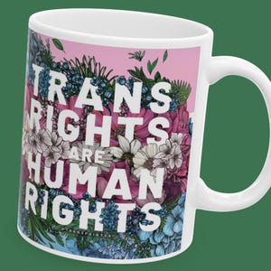 May include: White ceramic mug with a pink and blue floral design. The text "Trans Rights Are Human Rights" is printed in white on the mug.