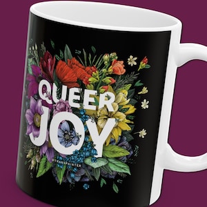 May include: Black coffee mug with a white design of a colorful floral bouquet and the text "QUEER JOY" in white letters. The text "TRANSPAINTER" is in smaller white letters below the bouquet.