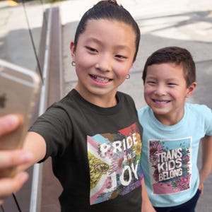 May include: Two children wearing t-shirts with text and floral designs. One shirt says "PRIDE AND JOY" with a rainbow flag design. The other shirt says "TRANS KIDS BELONG" with a floral border. The children are smiling and taking a selfie.