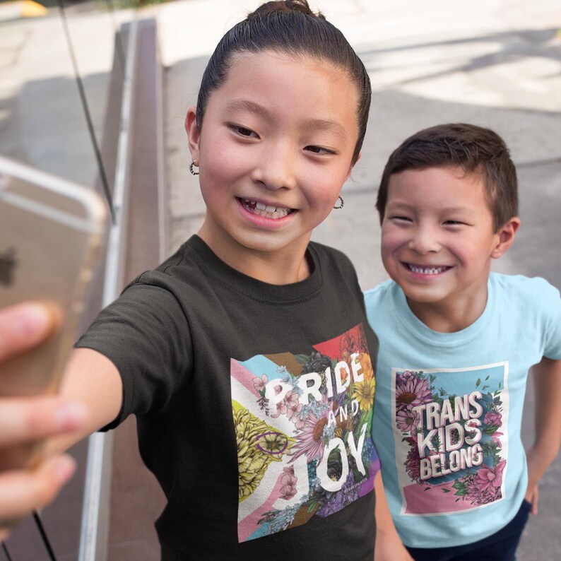 May include: Two children wearing t-shirts with text and floral designs. One shirt says "PRIDE AND JOY" with a rainbow flag design. The other shirt says "TRANS KIDS BELONG" with a floral border. The children are smiling and taking a selfie.