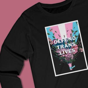 May include: Black sweatshirt with a graphic that reads "DEFEND TRANS LIVES." The graphic features the transgender pride flag colors, flowers, and leaves. The design is in a white rectangular frame.