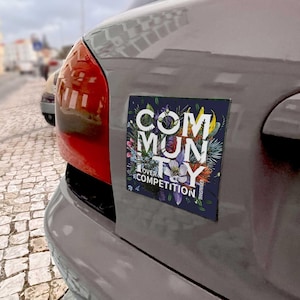 May include: A rectangular car magnet with the words "COMMUNITY OVER COMPETITION" in white, surrounded by a floral design. The magnet is affixed to the rear of a gray car, with a red taillight visible.