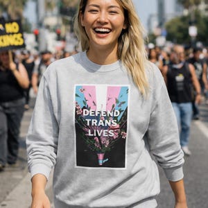 May include: A pale grey sweatshirt with the text "DEFEND TRANS LIVES". The graphic includes pink, blue, and white stripes with floral details. The sweatshirt is worn by a person smiling in an outdoor setting.