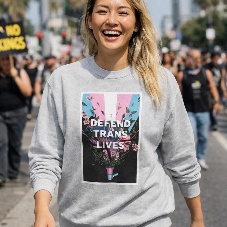 May include: A pale grey sweatshirt with the text "DEFEND TRANS LIVES". The graphic includes pink, blue, and white stripes with floral details. The sweatshirt is worn by a person smiling in an outdoor setting.