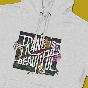 Puede incluir: Una sudadera con capucha gris con un gráfico cuadrado negro que presenta un diseño floral y el texto "Trans is Beautiful".