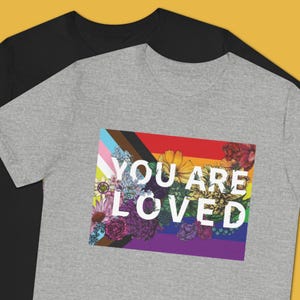 May include: Two t-shirts, one black and one gray, are displayed. The gray shirt features a colorful graphic with the text "YOU ARE LOVED" over a floral and rainbow design. The design incorporates various colors and floral elements.