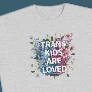 Peut inclure: Un t-shirt gris clair avec un motif floral et le texte "Trans Kids Are Loved" en lettres blanches. Les fleurs sont dans des tons de bleu et de rose.