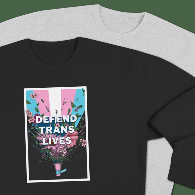 May include: Black sweatshirt featuring the text "DEFEND TRANS LIVES" above a floral design in pink, blue, and white. A grey sweatshirt is visible in the background.