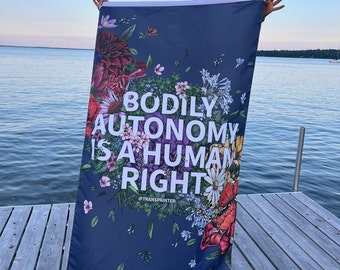 May include: A blue flag with a floral design and the text "Bodily Autonomy is a Human Right" in white lettering. The flag is held by a person on a wooden dock.