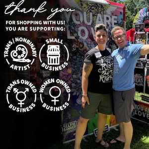 May include: Two people stand in front of a booth with a sign that says "You Are Loved". The sign is decorated with flowers and other colorful designs. The booth is selling items that support small businesses owned by women, transgender people, and non-binary artists. The sign also says "Thank You for Shopping with Us! You Are Supporting:"