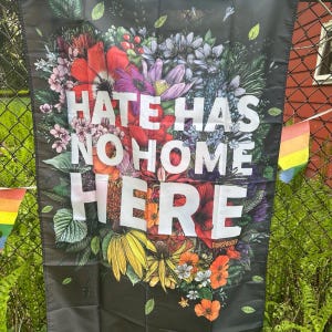 May include: A black flag with the words "HATE HAS NO HOME HERE" in large white letters, surrounded by a vibrant floral arrangement. The flowers include red, purple, yellow, and orange blooms. The word "TRANSPAINTER" is at the bottom.