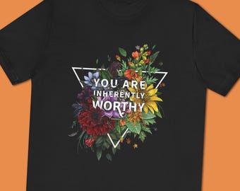 May include: Black t-shirt featuring a floral design within a white triangle. The design includes colorful flowers and the text "YOU ARE INHERENTLY WORTHY" in white. The shirt is a crew neck style.