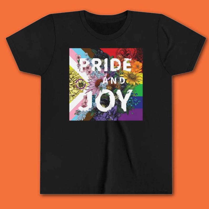 May include: Black t-shirt featuring a floral design with the words "Pride and Joy" in white. The floral arrangement incorporates colors from various pride flags, including the transgender flag, rainbow flag, and others.