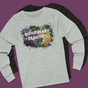 May include: A light gray long-sleeve t-shirt layered over a black long-sleeve t-shirt. The gray shirt features a graphic of colorful flowers in shades of purple, yellow, and green with the white text "NONBINARY IS BEAUTIFUL" printed in the center.