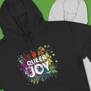 May include: Black hoodie with a floral design and the words "QUEER JOY" in white. The floral arrangement includes red, purple, yellow, and blue flowers. The hoodie has a drawstring hood and a gray hood peeking out from behind.