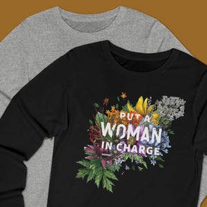 May include: Two long-sleeved shirts, one gray and one black, are displayed. The black shirt features a floral design with the text "PUT A WOMAN IN CHARGE" in white. The floral arrangement includes yellow, blue, and purple flowers.