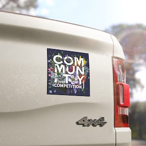 May include: A square magnet with the words "COMMUNITY OVER COMPETITION" in white, set against a dark blue background with a floral design. The magnet is affixed to the back of a white vehicle.