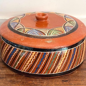 May include: A round, terracotta-colored ceramic casserole dish with a lid and handle. The lid and body are decorated with colorful geometric patterns in blue, green, yellow, and black. The dish sits on a wooden surface.