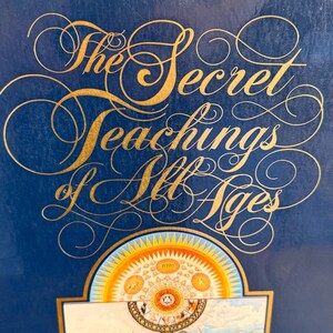Może przedstawiać: Książka zatytułowana "The Secret Teachings of All Ages" autorstwa Manly'ego P. Halla. Okładka jest granatowa ze złotymi literami i elementami dekoracyjnymi. Na dole znajduje się kolorowa ilustracja.