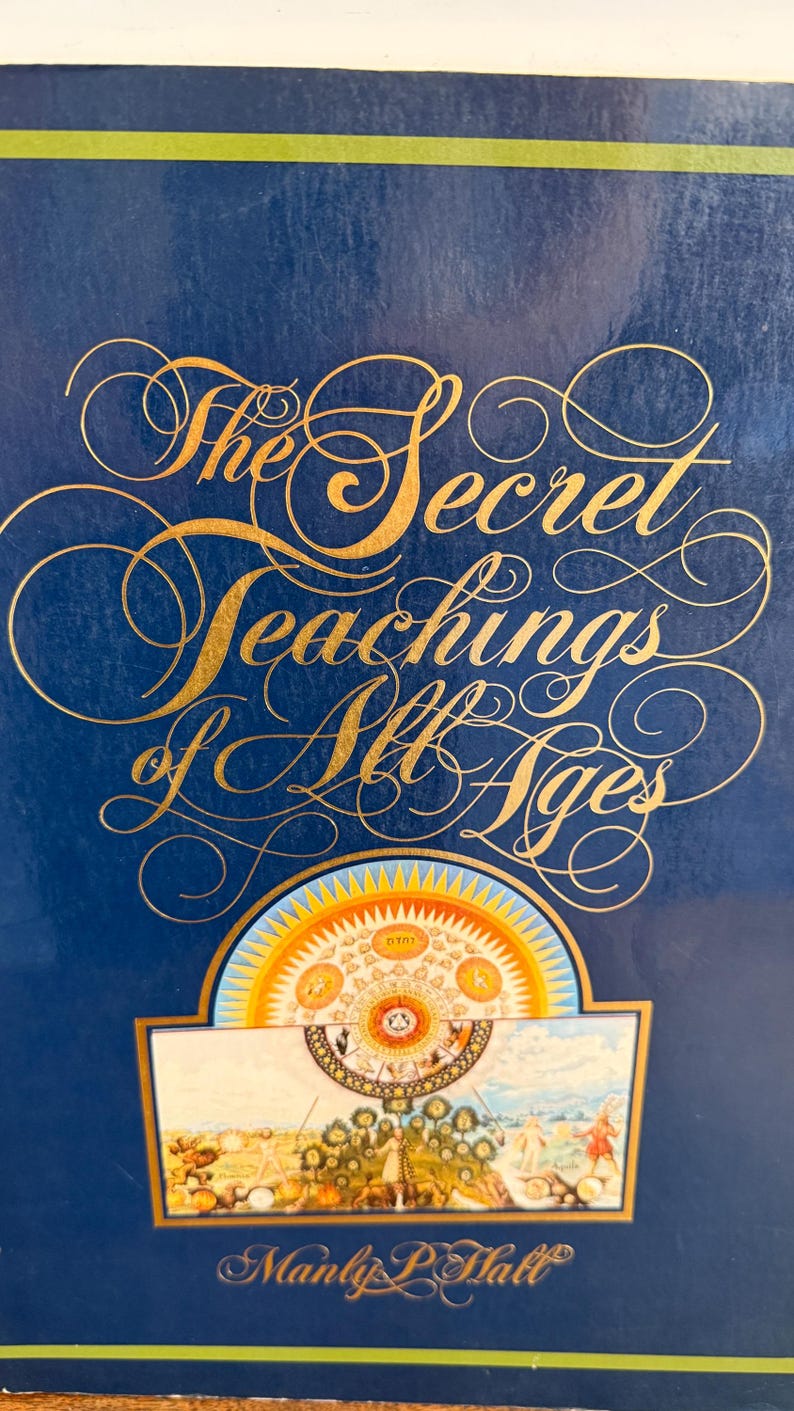 Może przedstawiać: Książka zatytułowana "The Secret Teachings of All Ages" autorstwa Manly'ego P. Halla. Okładka jest granatowa ze złotymi literami i elementami dekoracyjnymi. Na dole znajduje się kolorowa ilustracja.