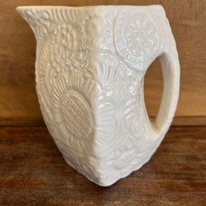 May include: White ceramic pitcher with a floral embossed design. The pitcher has a wide mouth and a handle.