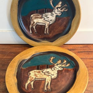 May include: Two round ceramic plates featuring a central embossed reindeer design. The plates have a wide tan rim, a dark brown lower section, and a teal blue upper section. The reindeer is depicted in a light cream color with dark outlines.