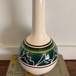 May include: A cream-colored ceramic vase with a long neck and a rounded base. The base features a teal, green, and black geometric design. The vase is resting on a vintage book.