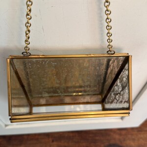 May include: A rectangular, hanging glass terrarium with a brass frame and chain. The glass has a slightly aged appearance. The terrarium is suspended from a brass chain, suggesting it can be hung as a decorative item.