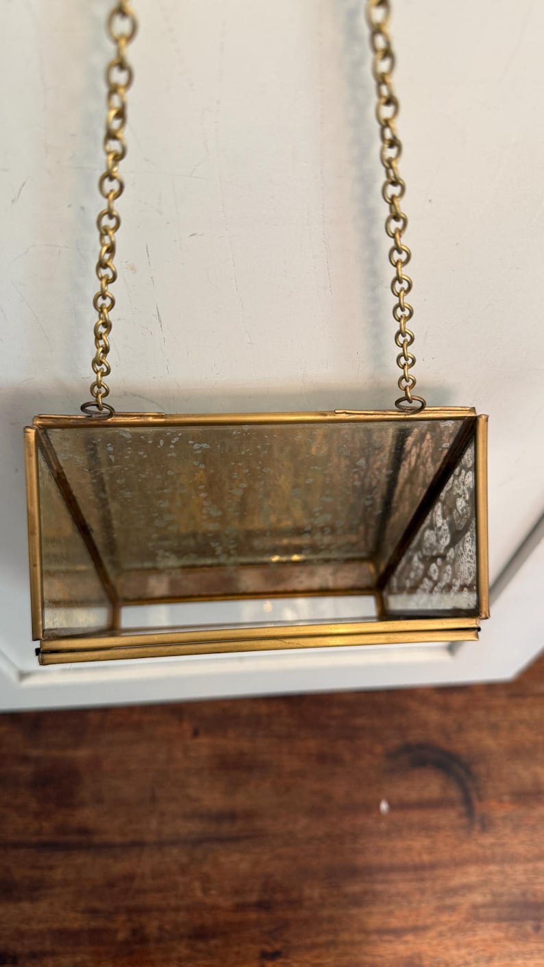 May include: A rectangular, hanging glass terrarium with a brass frame and chain. The glass has a slightly aged appearance. The terrarium is suspended from a brass chain, suggesting it can be hung as a decorative item.