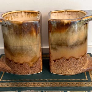 May include: Two ceramic mugs with a unique rectangular shape and integrated handles. The mugs feature a gradient of brown, tan, and beige tones, with a textured base. The design is reminiscent of vintage or mid-century modern pottery.